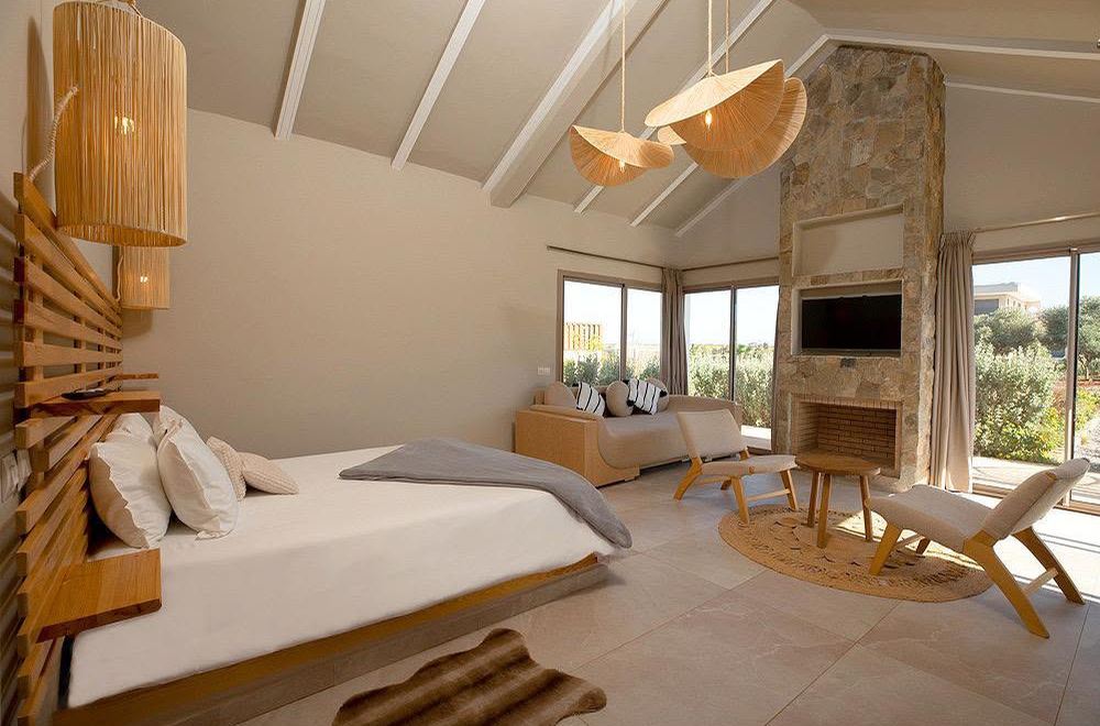 Chambre lumineuse sous charpente, cheminée en pierre et fauteuils en bois clair