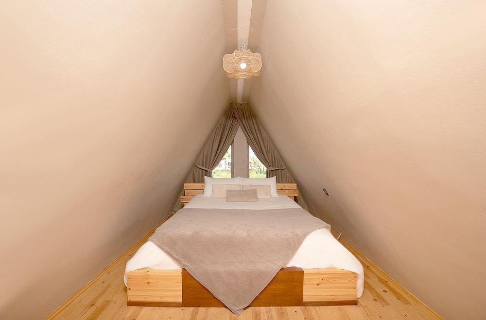 Chambre mansardée sous charpente triangulaire, lit en bois clair face à la fenêtre
